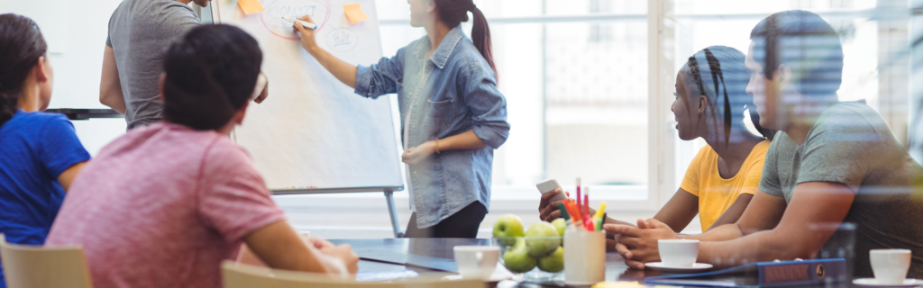 Meeting with 6 colleagues, female is writing on a whiteboard sitting in a corporate office