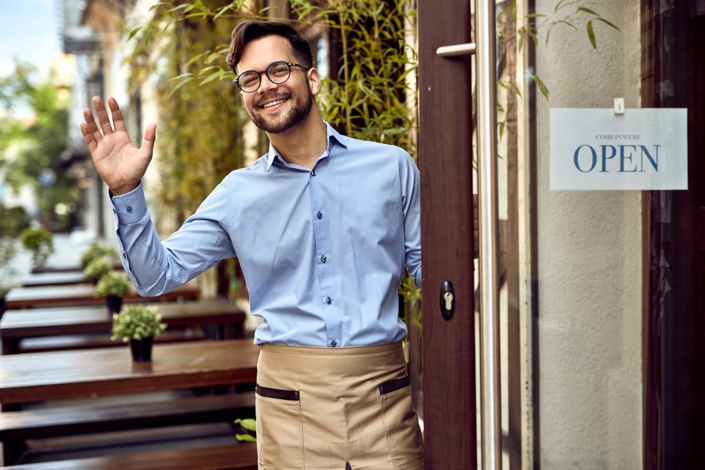 Young happy waiter standing at cafe's front door and waving to someone.